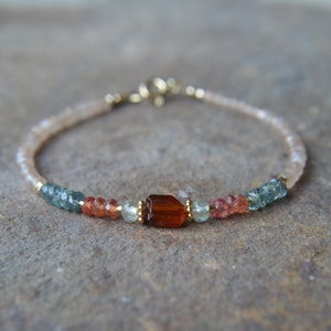 May include: A delicate bracelet with a mix of light pink, orange, and blue gemstones. A larger, rectangular, brown gemstone is the centerpiece of the bracelet.