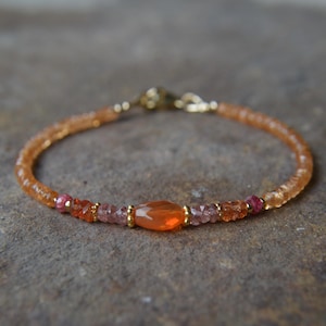 May include: A delicate bracelet featuring small, faceted orange and pink gemstone beads. A larger, central faceted orange stone is flanked by smaller pink and orange beads, accented with tiny gold-toned metal spacers and a gold-toned lobster clasp.