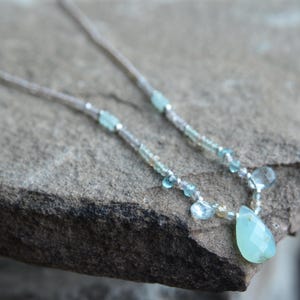May include: Delicate necklace featuring a light blue, teardrop-shaped pendant. The necklace is strung with small, light blue, and clear beads. The necklace is displayed on a textured, gray stone surface.