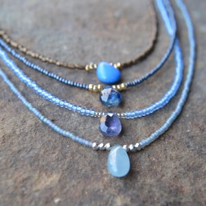 May include: Four delicate necklaces with gemstone pendants in shades of blue and purple. The necklaces feature small beads and metallic accents. The gemstones include a teardrop-shaped aquamarine, a faceted sapphire, and a smooth blue stone.