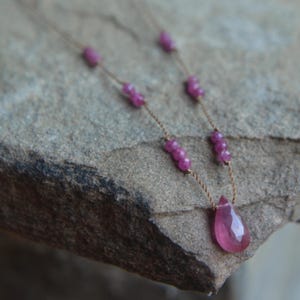 May include: A delicate necklace featuring a teardrop-shaped pink gemstone pendant. The necklace has a thin, gold-colored chain with small, round pink beads spaced along its length. The jewelry is displayed on a textured, gray stone surface.