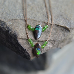 May include: Two layered necklaces with oval-shaped opal pendants. The opals display a mix of green, blue, and brown colors. Each necklace features small green beads and a brown cord. The necklaces are displayed on a textured gray stone.