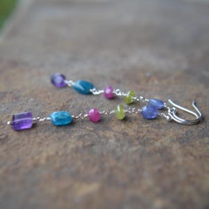 May include: Dangling earrings featuring a variety of colorful gemstones. The earrings have a silver chain with amethyst, turquoise, pink, green, and blue stones. The earrings are displayed on a brown surface.