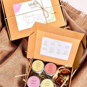 Könnte beinhalten: Zwei offene braune Geschenkboxen aus Pappe mit Tee-Artikeln. Eine Box enthält vier kleine Teedosen, ein Honigglas und ein Tee-Ei. Die andere Box ist geschlossen und mit Bindfaden verschnürt. Die Boxen stehen auf einer Jutefläche.