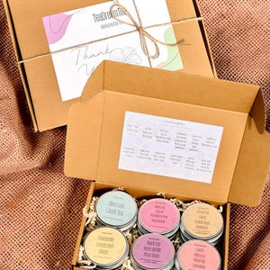 Könnte beinhalten: Ein Geschenkset mit Kräutertees. Die offene Box zeigt sechs kleine, runde Dosen mit farbenfrohen Etiketten, die jeweils eine andere Teemischung enthalten. Die Box ist mit geschreddertem Papier und einer Liste von Teesorten gefüllt. Der Deckel hat eine Dankeskarte.