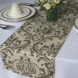 May include: A beige table runner with a brown damask pattern. The runner is on a white tablecloth and there is a small white cake and a crystal candle holder on top of it.