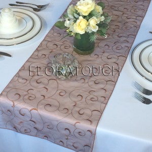 May include: A light brown table runner with a delicate swirl pattern embroidered in a darker brown. The runner is laid on a white tablecloth and features a vase of yellow roses and a clear glass candle holder.