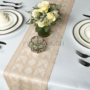 May include: A beige lace table runner with a geometric pattern. The runner is laid on a white tablecloth and features a vase of yellow roses and a clear glass candle holder.