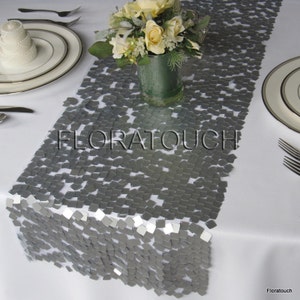 May include: Silver sequin table runner with a square pattern. The runner is on a white tablecloth with a vase of white flowers in the center.