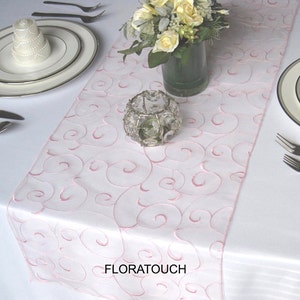 May include: A white table runner with a pink swirl design. The runner is made of a sheer fabric and has a delicate, feminine look. The text "FLORATOUCH" is printed on the runner.