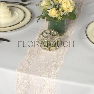 May include: A white table runner with a pink lace trim. A clear glass candle holder with a crystal design sits on the runner. A bouquet of yellow roses in a green vase is in the background.