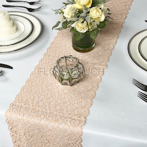 May include: A beige lace table runner with scalloped edges. The runner is laid on a white tablecloth and features a floral centrepiece in a clear glass vase.
