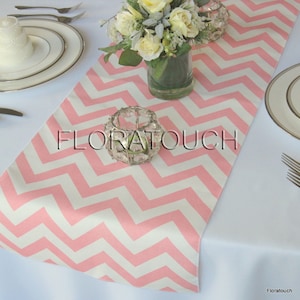 May include: Pink and white chevron table runner with the text "Floratouch" printed on it. The runner is on a white tablecloth with a vase of white flowers and a candle holder.