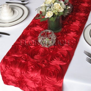 May include: Red satin table runner with a 3D rose design. The runner is on a white tablecloth and has a vase of white flowers and a candle holder on top.