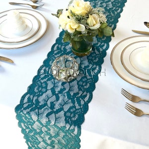 May include: Teal lace table runner with a floral design. The runner is on a white tablecloth with a vase of white flowers and a clear glass candle holder in the center.