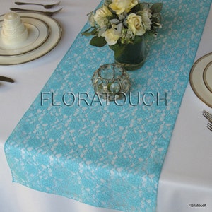 May include: A turquoise lace table runner with a floral design. The runner is on a white tablecloth with a vase of white roses and a clear glass candle holder.