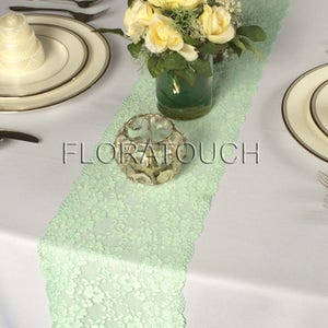 May include: A light green lace table runner with a clear glass votive candle holder on top. The table runner is on a white tablecloth with a white cake stand and a bouquet of yellow roses in a green vase.