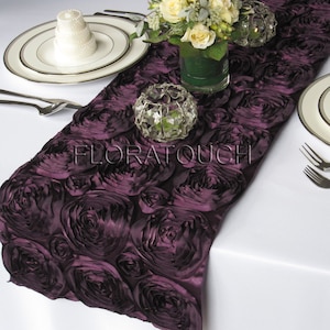 Peut inclure: Un chemin de table en satin violet avec un motif de roses 3D. Le chemin de table est sur une nappe blanche avec un support à gâteau blanc et un vase de fleurs blanches.