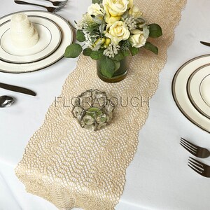 May include: A beige lace table runner with a delicate floral pattern. A clear glass candle holder with a tealight is placed on the runner. A bouquet of white roses is in a clear glass vase on the runner.