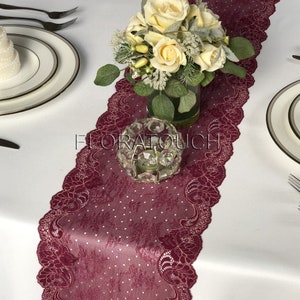 May include: Burgundy lace table runner with a delicate floral pattern and white polka dots. The runner is 12 inches wide and features a scalloped edge. A clear glass vase with white roses and greenery sits on the runner.