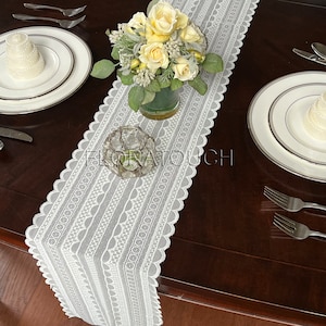 May include: White lace table runner with a scalloped edge and a repeating pattern of circles and arches. A bouquet of yellow roses in a glass vase is centered on the runner.