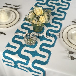 May include: A white table runner with a teal geometric pattern. The runner is decorated with a small bouquet of yellow flowers in a clear glass vase and a crystal candle holder.