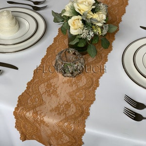 May include: A gold lace table runner with a scalloped edge. The runner is on a white tablecloth with a bouquet of yellow roses in a clear vase and a silver candle holder.
