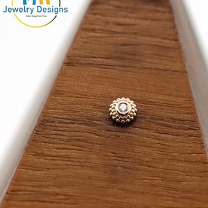 Könnte beinhalten: Ein kleiner, goldfarbener Ohrstecker mit einem zentralen, klaren Edelstein, umgeben von einem Perlendesign. Der Ohrring ist auf einer Holzoberfläche platziert. Das Logo "14k Jewelry Designs" ist sichtbar.