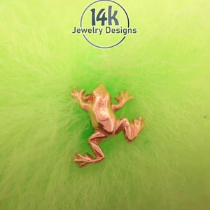 May include: A small, metallic, rose-gold colored frog-shaped jewelry piece. Above the frog, a circular logo contains the text 14k Jewelry Designs. The frog is depicted in a jumping position with its limbs extended outward from its body.