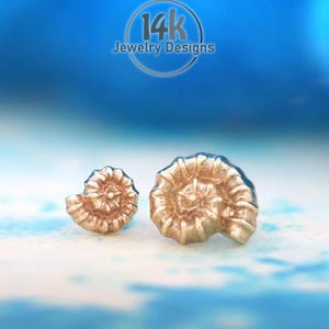 May include: Two gold-toned stud earrings shaped like ammonite fossils with a spiral shell pattern. The text 14k Jewelry Designs is visible in a circular logo at the top center of the image. The earrings have a metallic finish and texture.