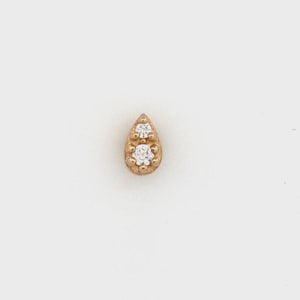 May include: A gold-colored teardrop-shaped earring with two clear, round gemstones. The earring has a textured surface and is set against a white background. The gemstones are positioned near the top and center of the earring.
