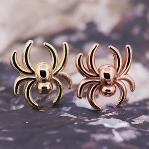 May include: Two spider-shaped stud earrings. One is gold-colored, the other is rose gold. Each earring has a rounded body and eight curved legs. The earrings are displayed on a textured, blurred background.
