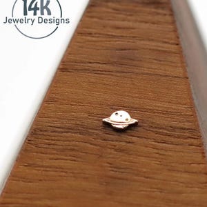 May include: A small, gold-colored planet-shaped stud earring. The earring has a circular body with a raised, ring-like band around the center. The earring is on a wooden surface. The image includes the text "14k Jewelry Designs".
