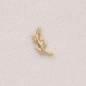Pode incluir: Um broche de metal dourado em forma de pequeno ramo fino com várias folhas delicadas que se estendem a partir do caule central. A peça apresenta uma superfície lisa e polida com um design curvo que termina em ponta na extremidade inferior.