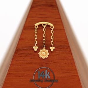 Könnte beinhalten: Ein goldfarbenes Schmuckstück mit floralem Design und herabhängenden Ketten. Das Stück ist auf einer Holzoberfläche platziert und zeigt den Text "14k Jewelry Designs" in einem kreisförmigen Logo.