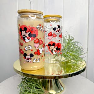 May include: Two clear glass tumblers with bamboo lids and red straws. The tumblers feature a Valentine's Day theme with Mickey Mouse and Minnie Mouse kissing and hearts.