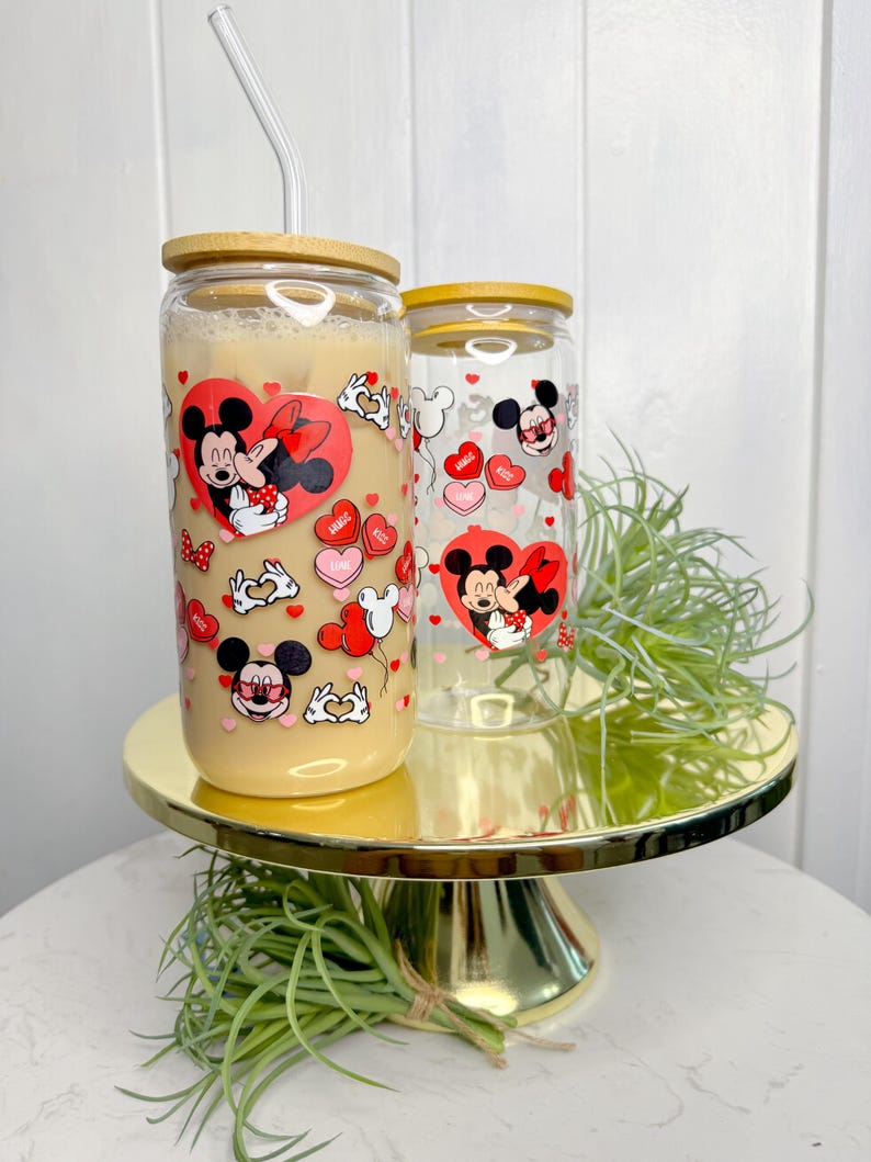 May include: Two clear glass tumblers with bamboo lids and red straws. The tumblers feature a Valentine's Day theme with Mickey Mouse and Minnie Mouse kissing and hearts.