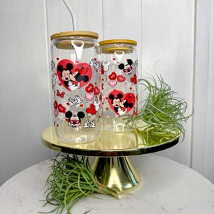 May include: Two clear glass tumblers with bamboo lids and straws. The tumblers feature a Valentine's Day design with Mickey Mouse and Minnie Mouse in a romantic embrace. The design includes red hearts, pink hearts, and white polka dots.