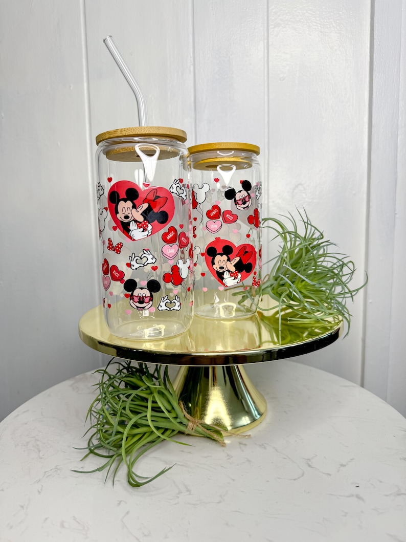 May include: Two clear glass tumblers with bamboo lids and straws. The tumblers feature a Valentine's Day design with Mickey Mouse and Minnie Mouse in a romantic embrace. The design includes red hearts, pink hearts, and white polka dots.