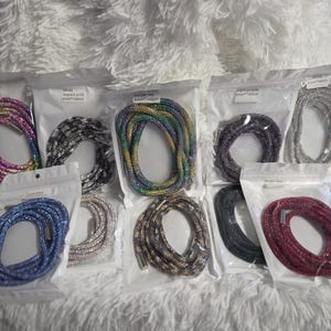 May include: A collection of ten individual rhinestone-encrusted cords, each packaged in a clear plastic bag. The cords feature various colours including rainbow, black, silver, blue, green, and red, with metallic end caps on each flexible strand.