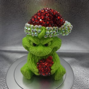 Puede incluir: Una figura verde de una criatura con el ceño fruncido, que lleva un gorro de Papá Noel cubierto de pedrería roja e iridiscente. La criatura sostiene en sus manos un pequeño adorno redondo incrustado con pedrería roja. Superficie texturizada.