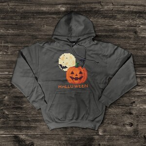 Puede incluir: Una sudadera con capucha gris oscuro con un diseño pixelado de Halloween. El diseño incluye una luna llena, una calabaza y la palabra "HALLOWEEN" en naranja. La sudadera tiene un bolsillo delantero y una capucha con cordón.