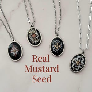 May include: Four silver-toned oval pendant necklaces with chain links. Each pendant features a black background with a unique white and gold-toned floral-inspired design containing a real mustard seed. The text "Real Mustard Seed" is centered.