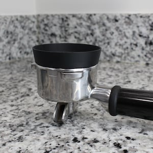 May include: A silver espresso portafilter with a black silicone tamper. The portafilter has a black handle.