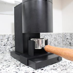 以下が含まれることがあります： ステンレス製のポルタフィルターと木製のハンドルが付いた黒いエスプレッソマシン。マシンはカウンタートップの上に置かれています。