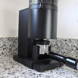 Puede incluir: Un molinillo de café negro mate con cuerpo cilíndrico y base rectangular vertical. Un portafiltros de metal plateado con mango negro está insertado en el soporte frontal. La parte superior cuenta con un dial con marcas numéricas blancas.