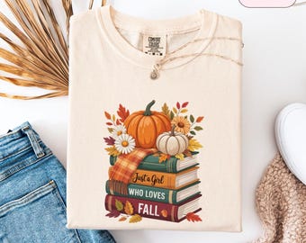 May include: Ivory-colored t-shirt with a fall-themed graphic. The design features a stack of books with the words "Just a Girl Who Loves Fall," pumpkins, fall leaves, and flowers. A gold necklace is draped across the shirt.