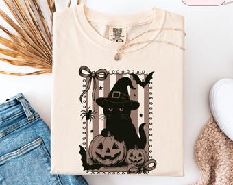 Könnte beinhalten: Elfenbeinfarbenes Sweatshirt mit einem Halloween-Motiv. Das Design zeigt eine schwarze Katze mit Hexenhut, Kürbissen, Fledermäusen und Spinnen. Der Text "IVORY" befindet sich oben rechts. Das Sweatshirt liegt gefaltet auf einer weißen Oberfläche.