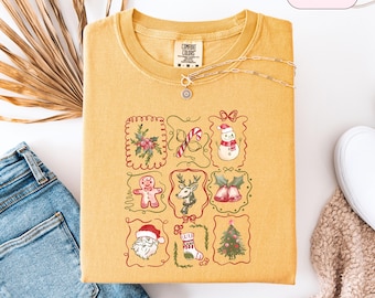 May include: Mustard-coloured sweatshirt with a crew neck and a festive Christmas-themed graphic. The graphic features nine framed illustrations including a snowman, Santa Claus, a Christmas tree, and a gingerbread man. The shirt is paired with blue jeans and a necklace.