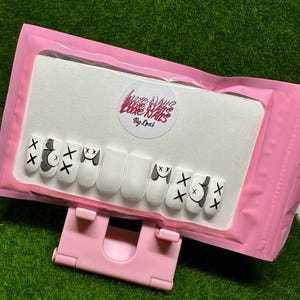 May include: A set of ten white press-on nails with a black and white cartoon design. The nails are displayed on a white card with the text "Love Nails Love Nails By Lexi".